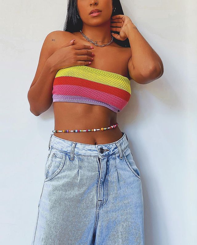 cinto-corrente-anos-2000-brendapabreu Garota usando top faixa de listras em amarelo, vermelho, rosa e lilás com calça jeans clara de cintura baixa e cinto de miçangas na barriga. Uma mão está acima do top e a outra está tocando a nuca.
