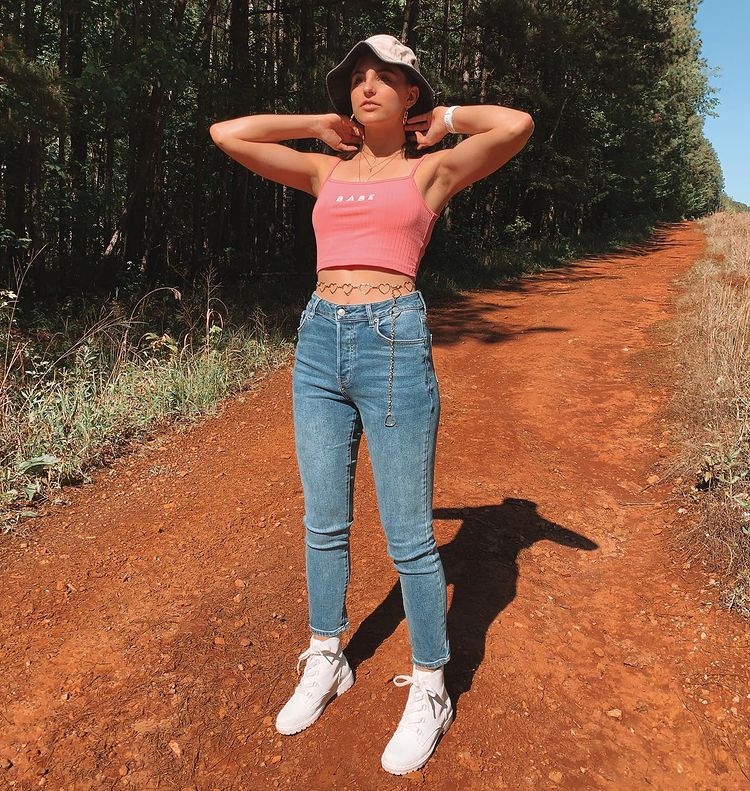 cinto-corrente-anos-2000-chloe5brooke Garota usando top cropped rosa, bucket hat, calça jeans skinny e coturno branco, além de cinto de correntes em formato de coração na barriga. Ela está em pé com os dois braços dobrados perto da nuca.