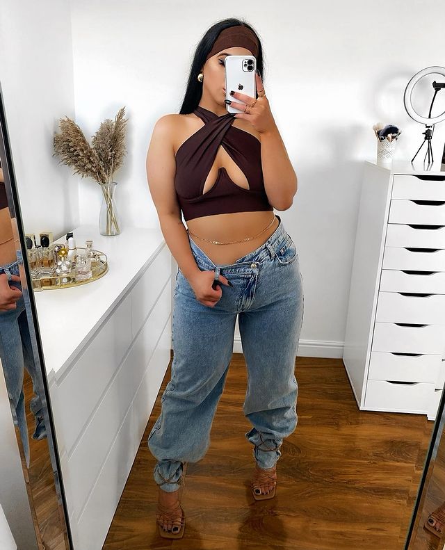 cinto-corrente-anos-2000-estefaniaa.jardim Mulher usando top cropped marrom com recorte no colo, calça jeans baggy e sandália marrom de tiras com bico quadrado. Ela também usa um cinto fino dourado na barriga, está em pé em frente ao espelho, com o celular na frente do rosto, e uma mão segurando no passador da calça.