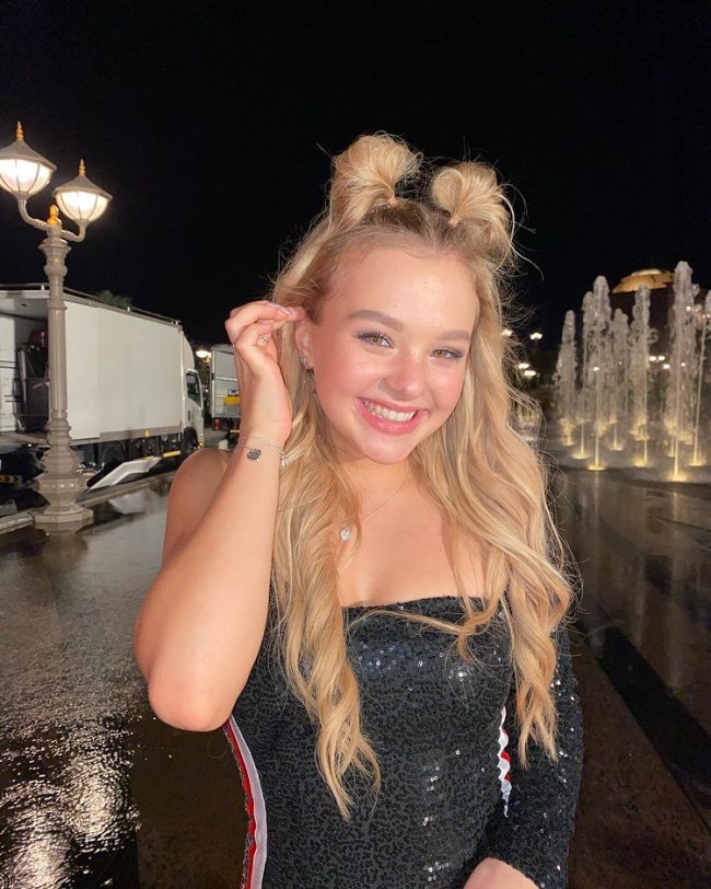 Sofya do Now United com coquinhos no cabelo Sofya Plotnikova, do grupo Now United, usando blusa preta tomara que caia com uma manga única, listras laterais, vermelha e branca, posando com uma mão mexendo no cabelo e usando pulseira prata.