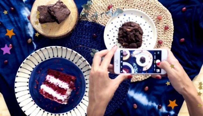 Mesa com toalha azul, com pratos, e bolos, sendo eles: red velvet, cupcake de chocolate e brownie. Também conseguimos ver duas mãos segurando um celular posicionado para fotografar a composição da mesa.