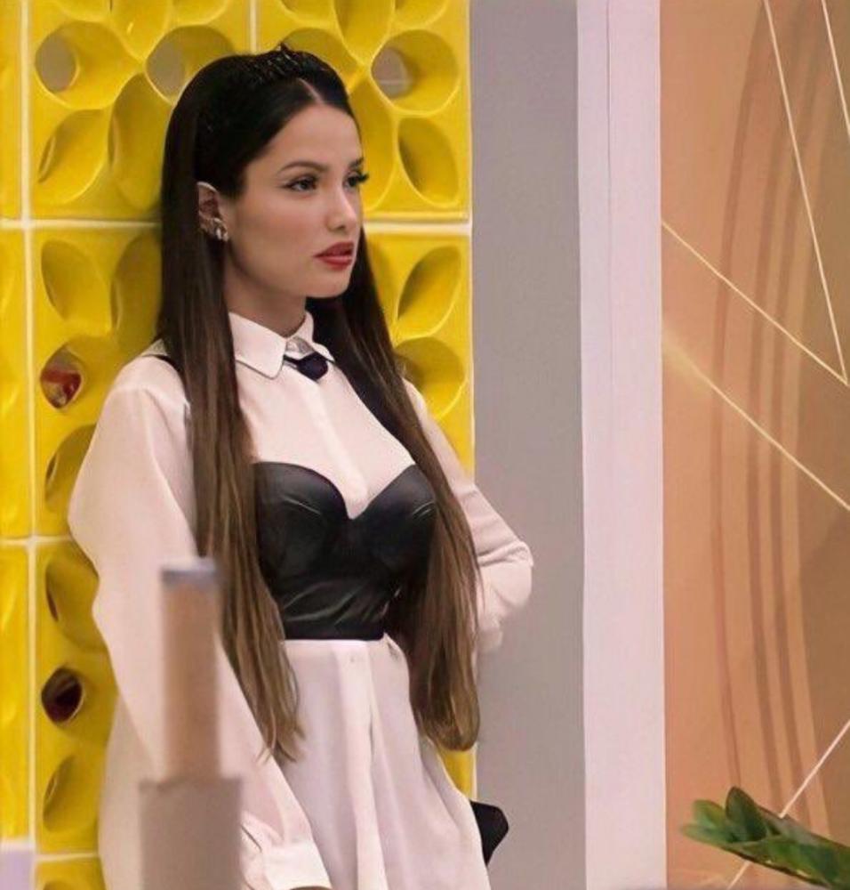 Juliette Freire no Big Brother Brasil 21 usando camisa branca, corset preto e tiara preta. Ela está encostada em uma parede amarela, com expressão facial séria, e a foto foi tirada de lado.