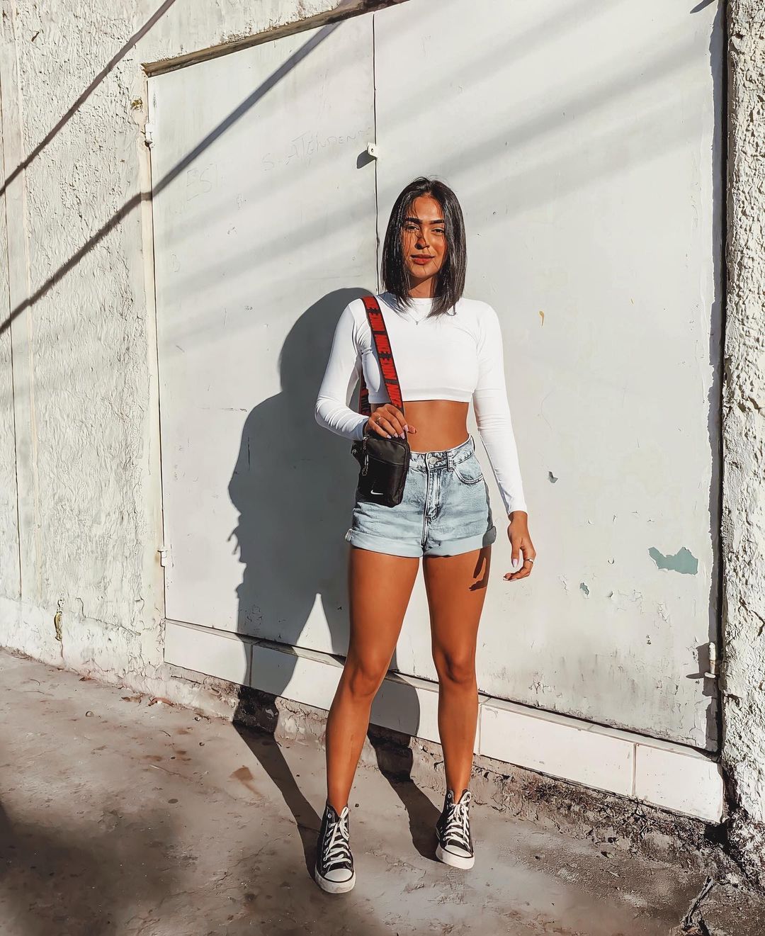 Garota usando top cropped branco de manga longa com short jeans e tênis All Star preto e branco de cano alto. Em pé, ela está segurando uma minibolsa de alça longa em um dos ombros.