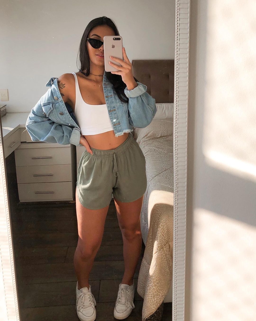 look-top-cropped-branco-dudaapaim Garota usando top cropped branco com jaqueta jeans, short de moletom verde e tênis branco. Ela está em pé, de frente para um espelho, tirando a foto com óculos de sol, com uma das mãos segurando o celular e, a outra, na cintura.
