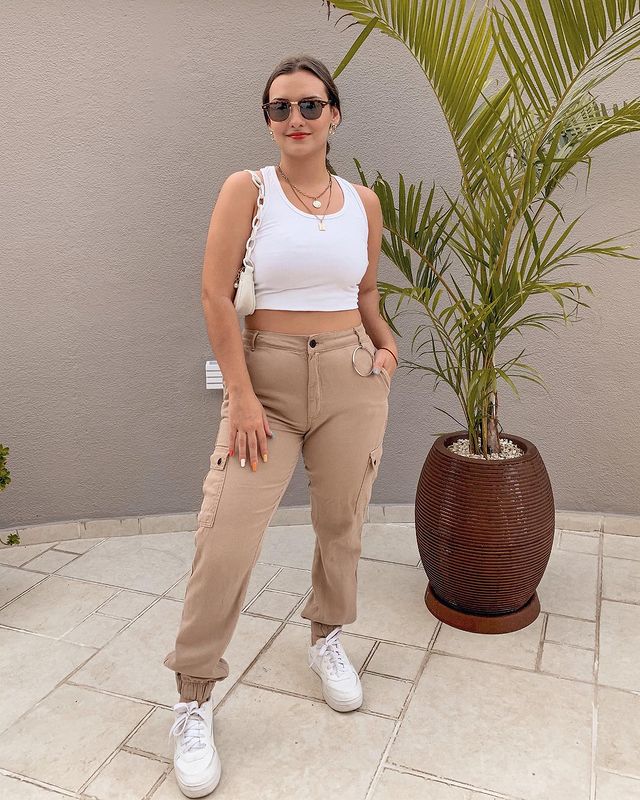 Garota usando top cropped branco com calça cargo bege e tênis branco. Ela está em pé, com uma bolsa branca pendurada em um dos ombros, óculos de sol e colares de correntes. Sua expressão facial tem um leve sorriso e uma das mãos está no bolso.