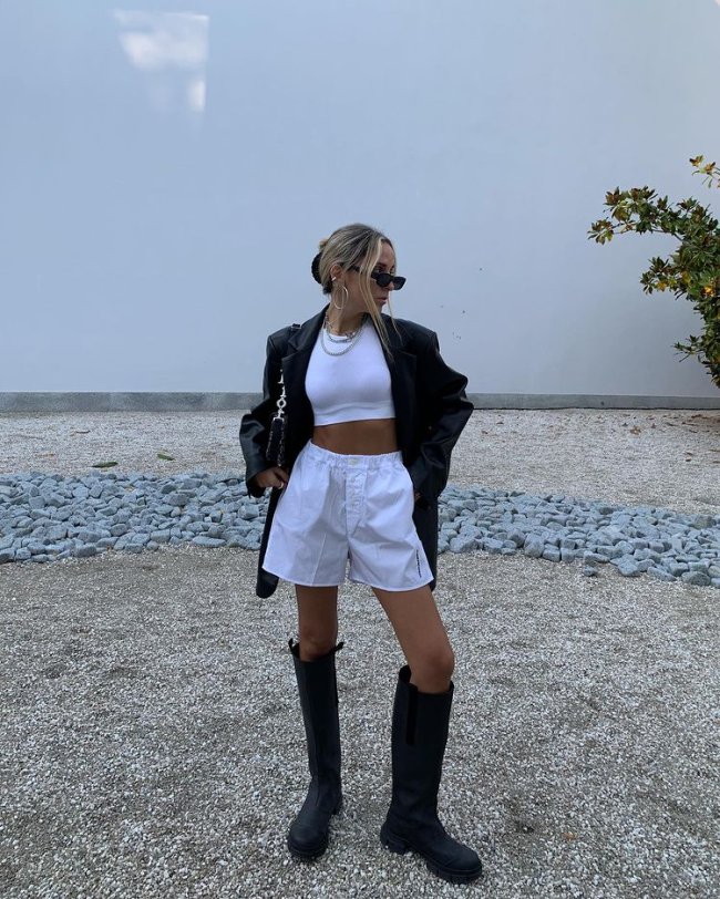 look-top-cropped-branco-sofiamcoelho Garota usando top cropped branco com short branco de cintura alta, bota tratorada preta que vai até o joelho e um casaco de couro preto por cima. Ela está em pé, usando óculos de sol e uma bolsa preta em um dos ombros. Sua cabeça está inclinada para um dos lados, e um dos joelhos está dobrado.