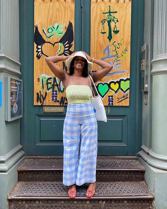 Garota usando top cropped sem alças verde com estampa floral e calça pantalona com estampa xadrez azul vichy. Ela ainda usa uma sandália vermelha, um chapéu bege e uma ecobag branca pendurada em um dos ombros. As duas mãos estão na cabeça e ela está sorrindo.