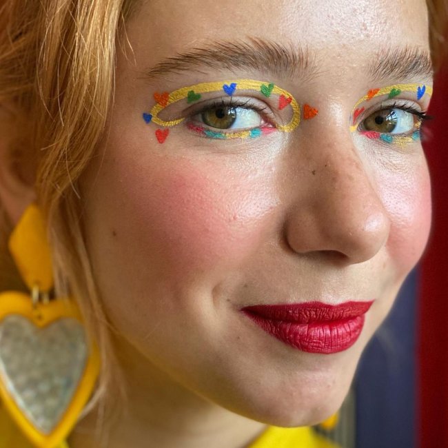 Jovem com o cabelo amarrado, brincos de coração amarelo, com maquiagem com sombra amarela e corações laranja e azul, nos lábios batom vermelho, olhando para a câmera com um quase sorriso.