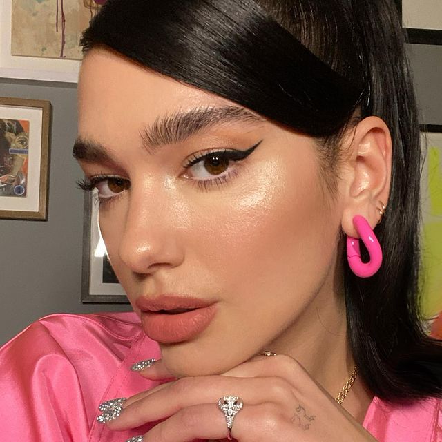 Maquiagem da Dua Lipa com delineado preto gatinho e pele iluminada. Na foto, aparece apenas o rosto dela. Ela está olhando de canto de olho, com uma expressão série e boca levemente aberta. Ela usa uma blusa e um brinco rosa neon.