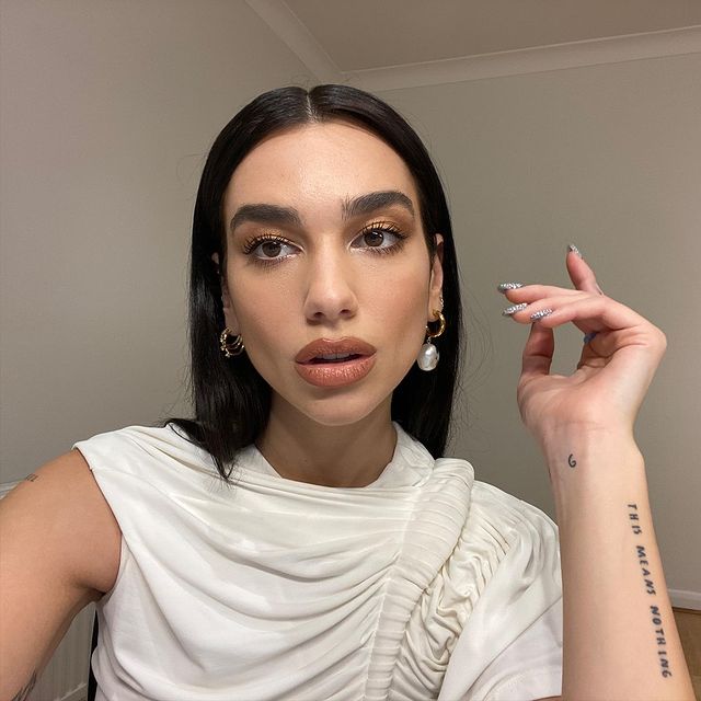 Dua Lipa usando maquiagem com sombra marrom esfumada. Ela está com uma blusa branca de um ombro só, uma das mãos levantadas e expressão facial séria com a boca levemente aberta.