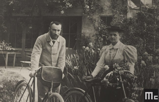 Radioactive: 10 coisas sobre Marie Curie que o filme da Netflix não conta Foto em preto e branco da cientista Marie Curie andando de bicicleta com seu marido, Pierre, em Paris; eles posam para a foto em uma vilinha florida