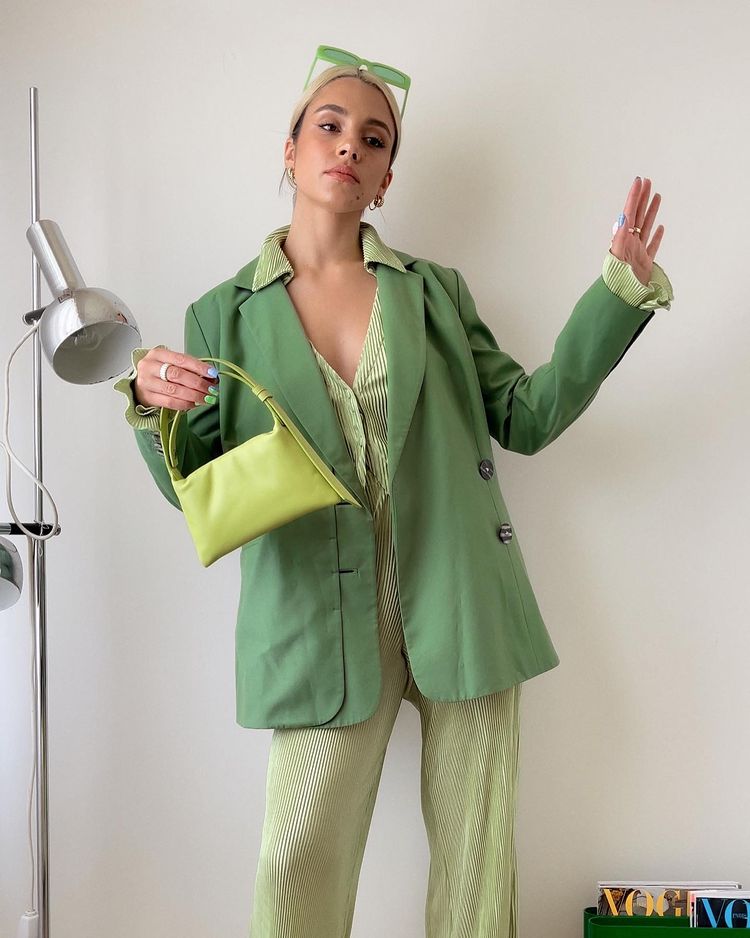 Garota usando look todo verde com blusa e calça listradas e blazer por cima. Com uma mão, ela segura uma bolsa pequena, enquanto a outra está levantada para cima com o braço levemente dobrado. Na cabeça, óculos de sol retangulares verdes.