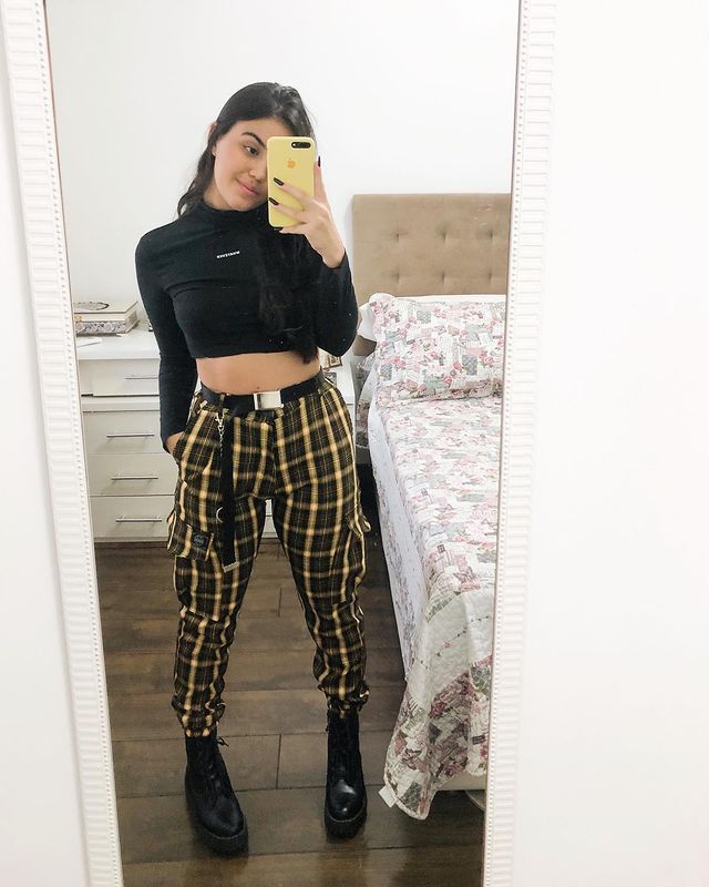 tendencia-outono-inverno-dudaapaim Garota usando blusa cropped preta de gola alta, calça xadrez preta e amarela e coturno preto. Ela está em pé em frente a um espelho com o celular cobrindo parcialmente o seu rosto.