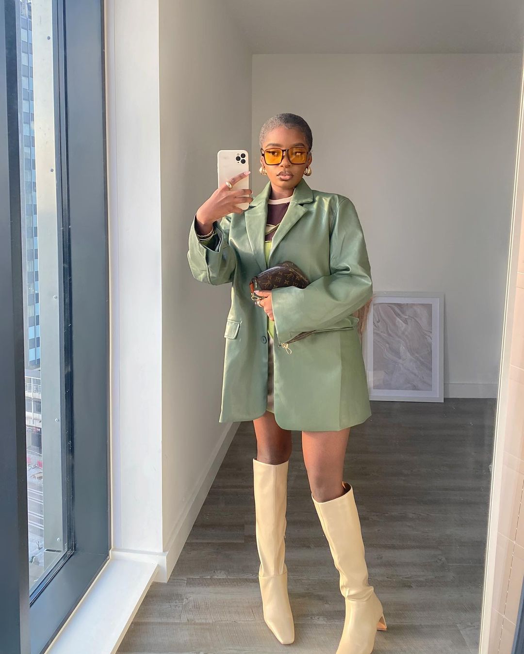 tendencia-outono-inverno-gratsikat Mulher usando casaco verde de couro com bota bege de bico quadrado que vai até o joelho e óculos de sol de lentes alaranjadas. Ela está em pé em frete a um espelho, segurando o celular com uma das mãos.