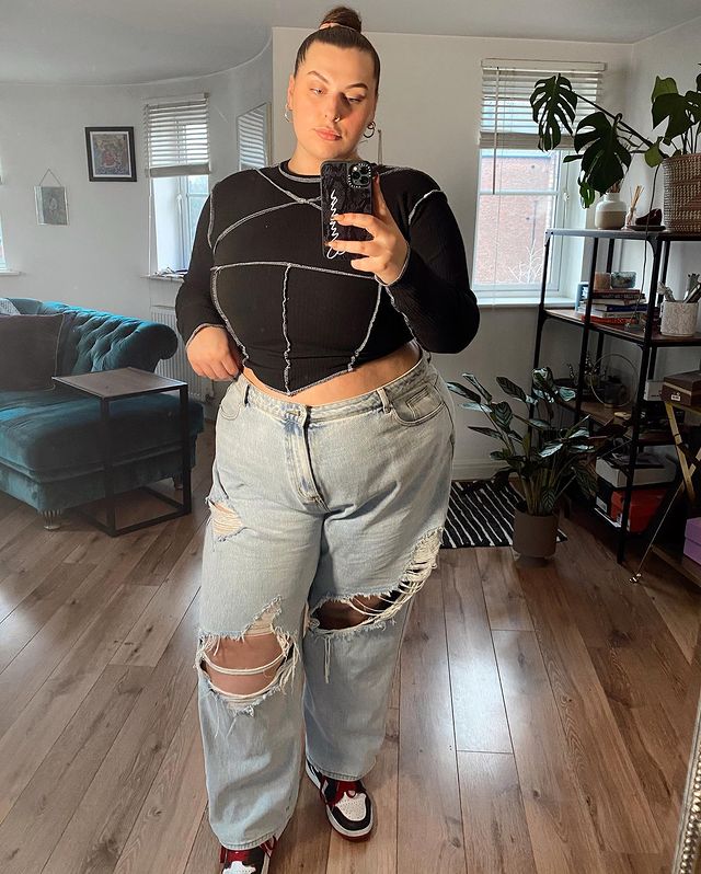 tendencia-patchwork-emmatamsinhill Garota usando blusa cropped preta de manga longa de patchwork com calça jeans rasgada no joelho e tênis branco, preto e vermelho de cano alto. Ela com expressão facial séria, em frente ao espelho, com uma das mãos na calça e a outra segurando o celular para tirar a foto.