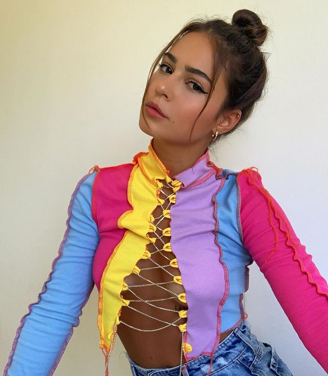 tendencia-patchwork-giubuscacio Garota usando blusa de patchwork amarela, lilás, azul e rosa com abertura no decote e detalhes de correntes prateadas. Ela está usando um coque alto com fios da franja na frente, cabeça levemente inclinada para o lado e expressão facial séria.