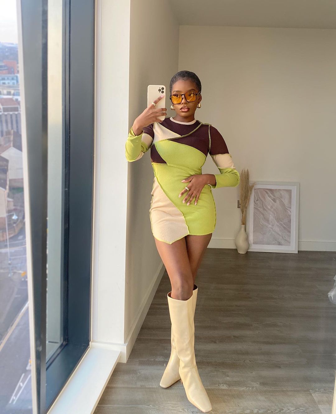 tendencia-patchwork-gratsikat Garota usando vestido de patchwork de manga longa nas cores verde neon, bege e marrom. Ela está com uma bota bege clara de bico quadrado que vai até o joelho e óculos com lentes alaranjadas. Ela está em pé, tirando a foto com o celular em uma das mãos, enquanto a outra está na cintura.