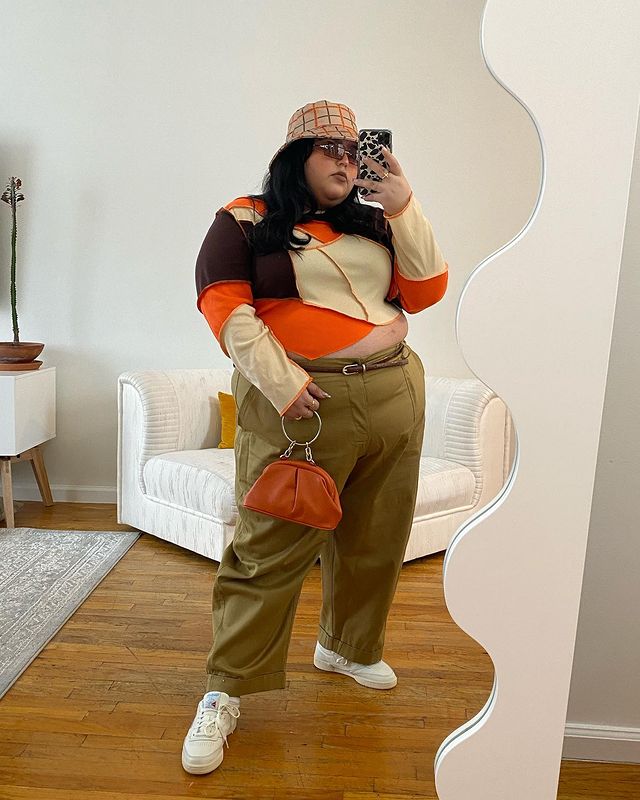 tendencia-patchwork-thisisjessicatorres Garota usando blusa de manga longa de patchwork nas cores laranja, bege e marrom. Ela está usando uma calça baggy bege escura, com cinto fino marrom, tênis branco e um bucket hat xadrez laranja e marrom. Ela está em pé, tirando foto em frente a um espelho com o celular na mão, e uma bolsa pequena laranja na outra, além de óculos de sol com lentes marrons.