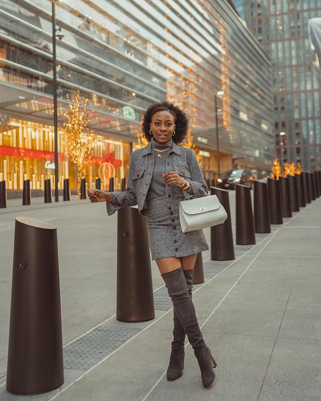 Garota está usando um look todo cinza com blusa de gola alta, casaco curto, saia de botões na frente e bota over the knee. Ela carrega uma bolsa pequena em um dos braços, e uma das pernas está cruzada na frente da outra.