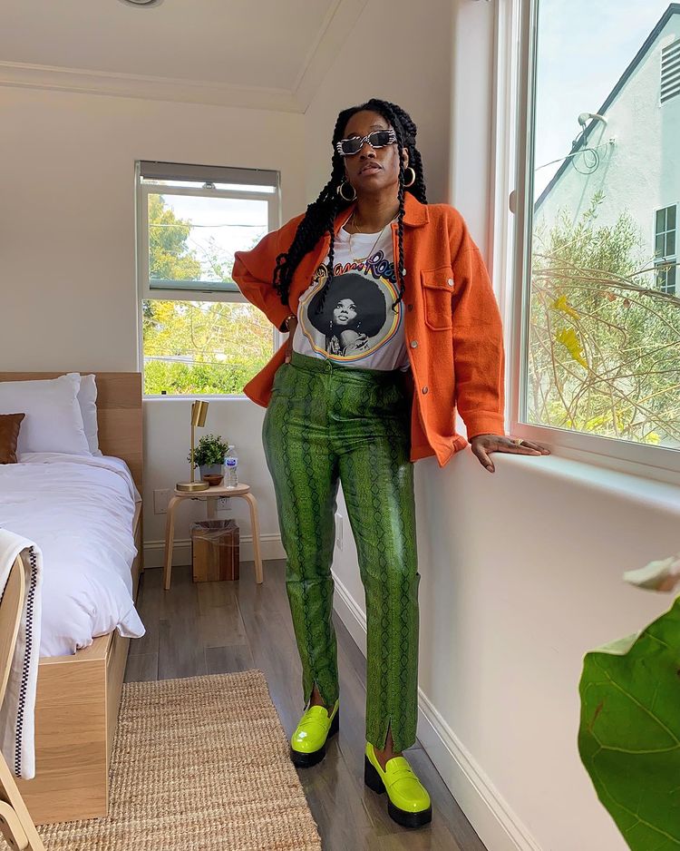 Mulher usando camiseta branca com casaco laranja, calça verde de estampa de cobra e oxford verde neon. Ela está com uma das mãos na cintura e a outra apoiada em uma janela.