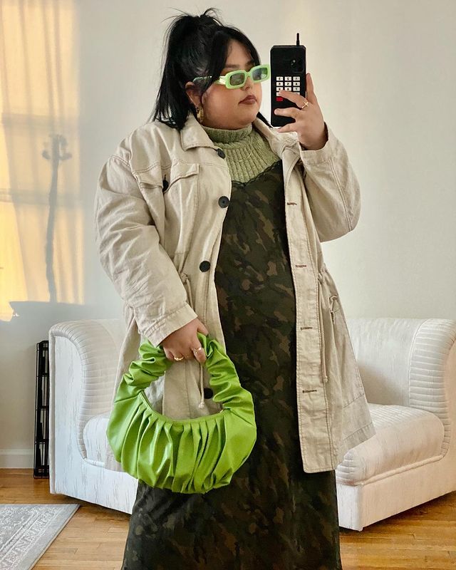 Mulher está usando uma blusa de gola alta verde com vestido verde escuro por cima, casaco bege de botões, bolsa e óculos de sol em verde neon. Ela está em frente ao espelho tirando a foto com o celular na mão.