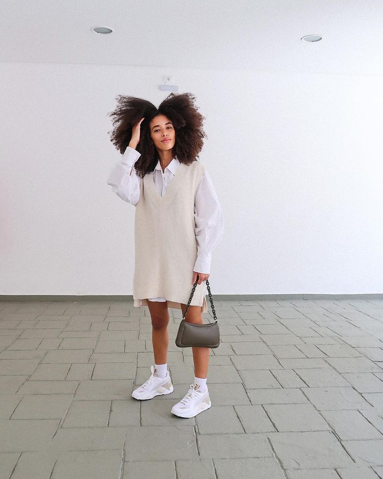 Garota usando camisa branca por baixo de colete bege oversized e tênis branco. Uma das mãos está segurando uma bolsa baguete cinza, e a outra está mexendo no cabelo crespo.