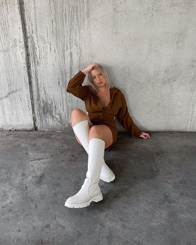 tendencias-outono-inverno-yourspruely-02 Mulher usando vestido marrom com mangas longas e bota até a altura do joelho off-white com sola tratorada. Ela está sentada no chão com uma mão na cabeça, a outra apoiada atrás e as pernas cruzadas.