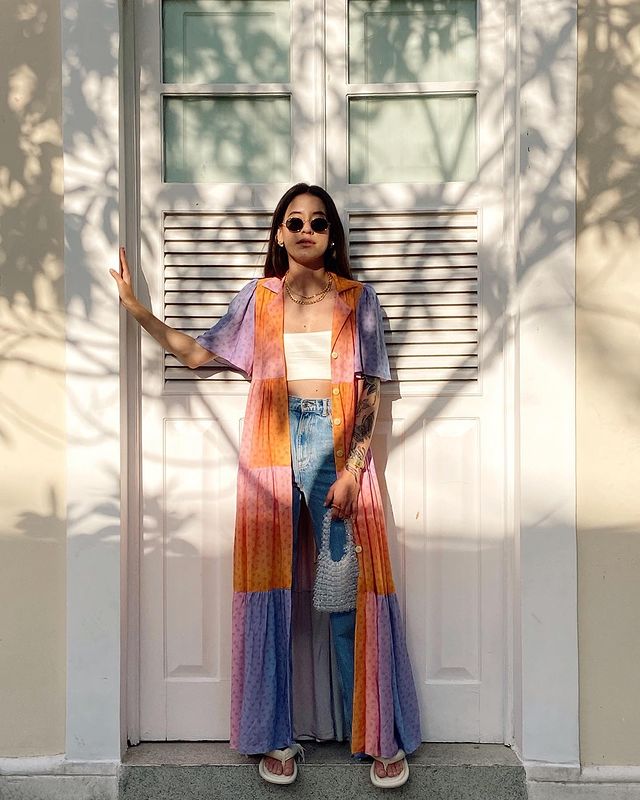 top-cropped-branco-schiavinis Garota usa top cropped branco com camisa longa de botões por cima nas cores laranja, rosa e lilás, uma calça jeans de cintura alta e um chinelo branco. Ela está apoiada em uma porta branca com uma das mãos, enquanto a outra segura uma bolsa branca de miçangas. Sua expressão facial é séria e ela usa óculos de sol redondos.