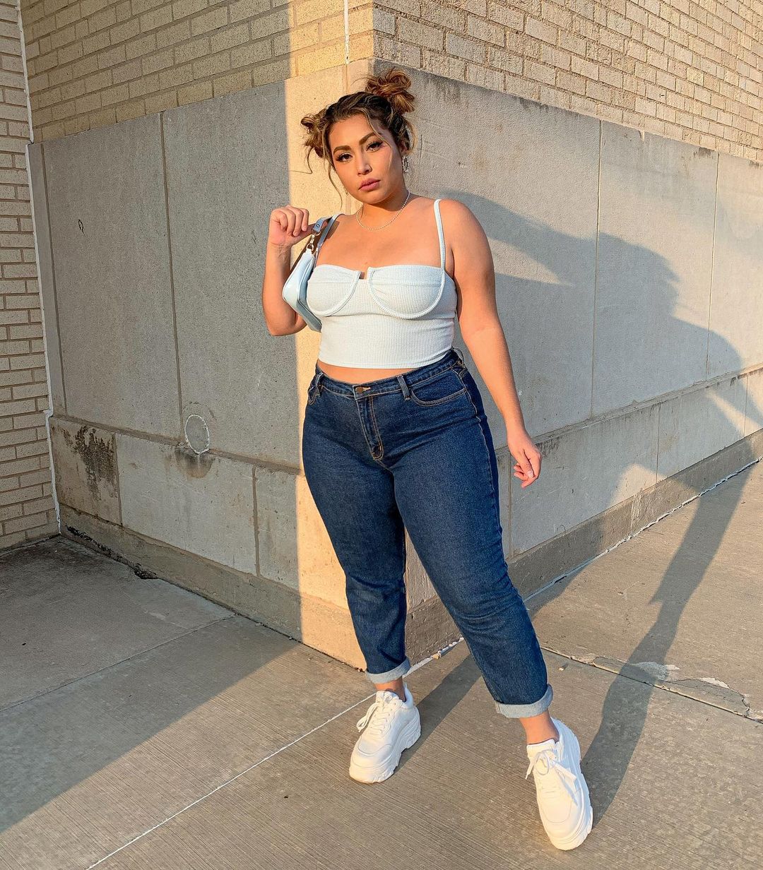 top-cropped-sutia-arlenett_ Garota usando top cropped branco com calça jeans de lavagem escura e tênis branco de paltaforma. Ela segura uma bolsa azul pastel de alça curta em um dos ombros. Sua expressão facial é séria e o cabelo está preso em dois coques no topo da cabeça.