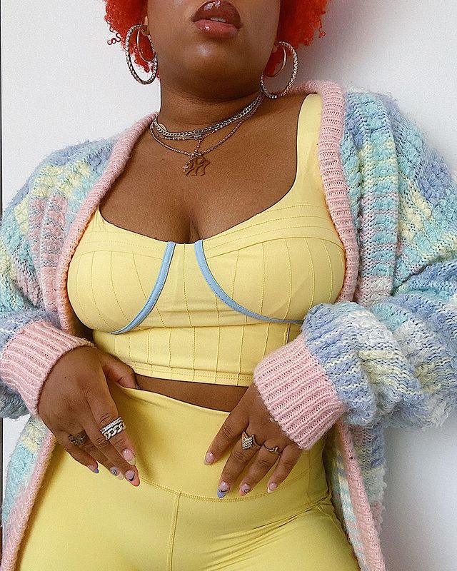 Garota usando top cropped amarelo com detalhes em azul no decote meia taça e legging da mesma cor. Por cima, um cardigan de tricô rosa e azul. Ela também usa vários anéis, colares e brincos em prata. Na foto, dá apenas para ver da boca para baixo até a altura do quadril.