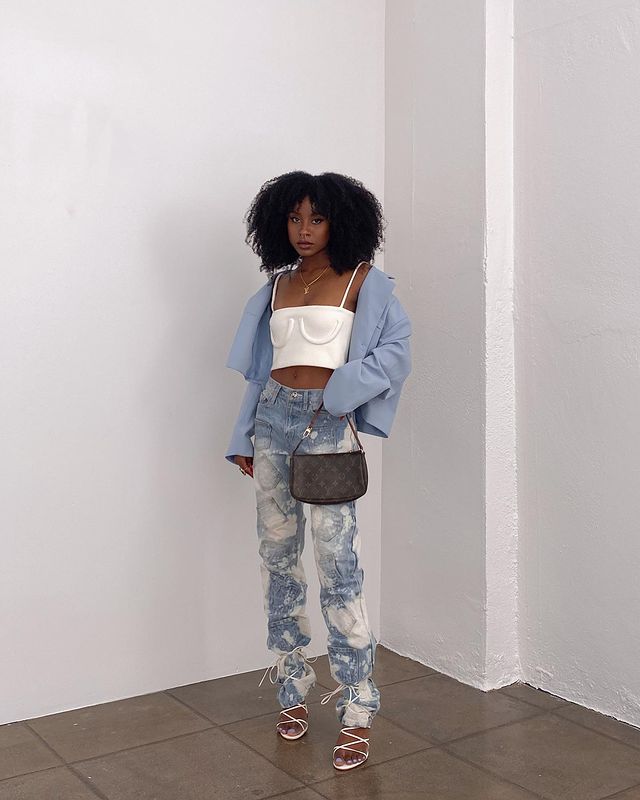 top-cropped-sutia-simply.cie Garota usando top cropped branco com jaqueta azul oversized, calça jeans tie-dye e sandália branca com amarrações por cima da calça. Ela está segurando uma bolsa da Louis Vuitton com uma das mãos e sua expressão facial é séria.