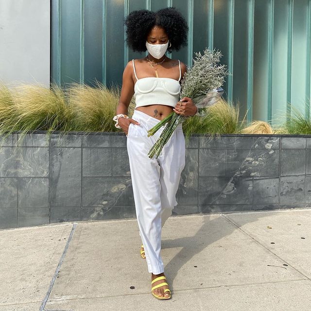 Garota usando top cropped off white com calça branca de tecido e rasteirinha amarela. Ela está usando uma máscara de proteção branca, olhando para baixo e segurando um buquê de flores com uma das mãos, enquanto a outra está no bolso.