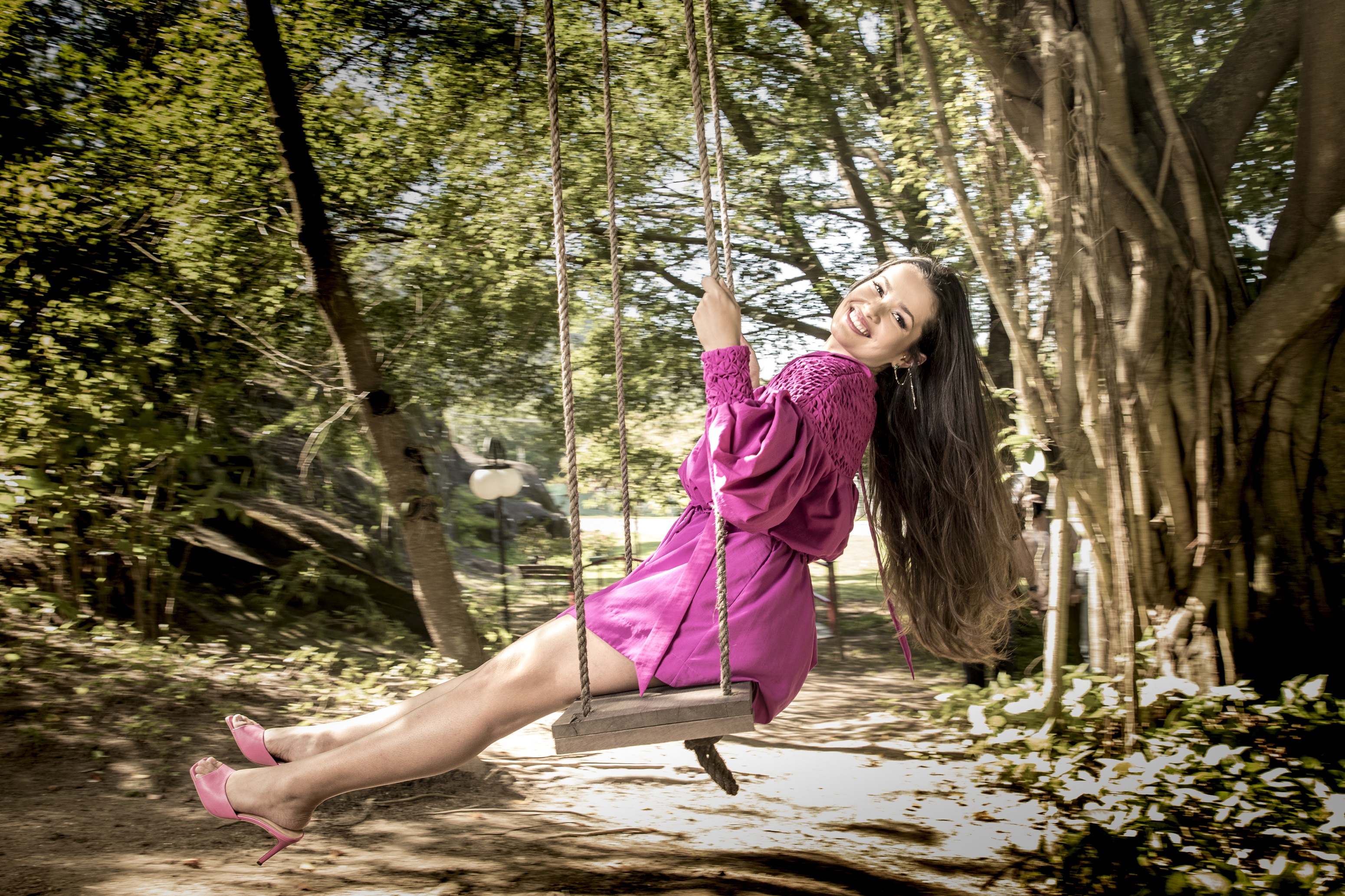 Juliette, vencedora do BBB21, em um balanço no meio de uma floresta usando um vestido rosa, sorrindo para câmera