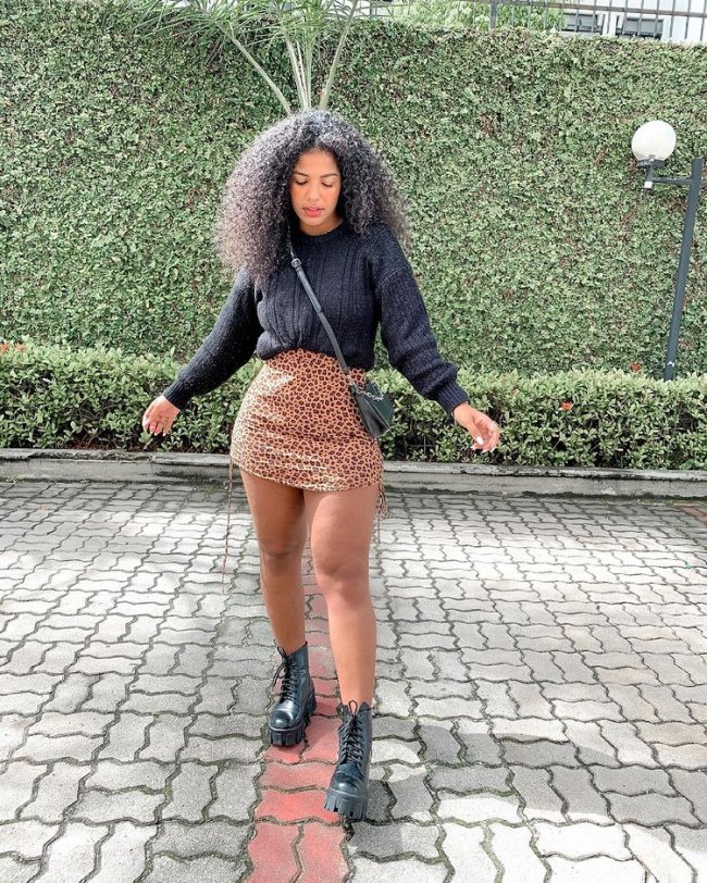 Bota coturno Jovem posando com os braços abertos, usando blusa preta, saia com estampa animal print, posando em frente a parede de folhas.