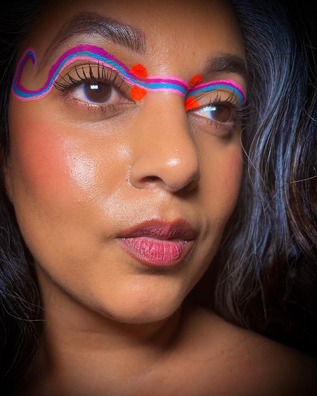 Delineado colorido unificado Foto com close no rosto de uma jovem com cabelo escuro, exibindo sua maquiagem com delineado unificado com traços rosa, azul e vermelho. Sua expressão é séria.