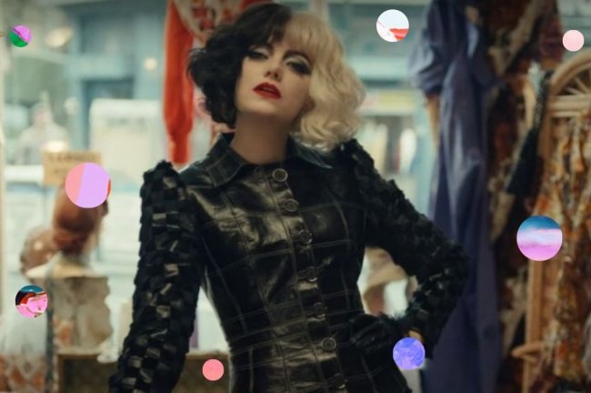 Emma Stone no filme Cruella Emma Stone no filme Cruella, vestida como a personagem, usando peruca de cabelo na cor preta e branca, jaqueta de couro preta, com a mão na cintura com expressão séria.