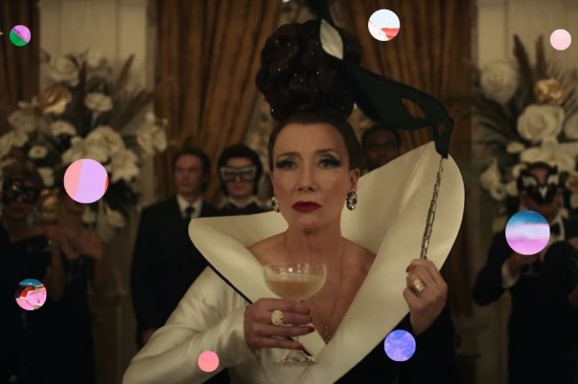 Emma Thompson como Baronesa Emma Thompson como Baronesa no filme Cruella, com roupa branca e preta, segurando uma mascara preta e uma taça na outra mão. Sua expressão é séria.