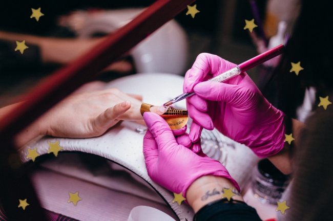Fazendo nail art Na imagem só conseguimos ver as mãos, de uma lado as mãos de uma profissional usando luva cor de rosa enquanto faz as unhas de uma outra pessoa que não conseguimos ver quem é.