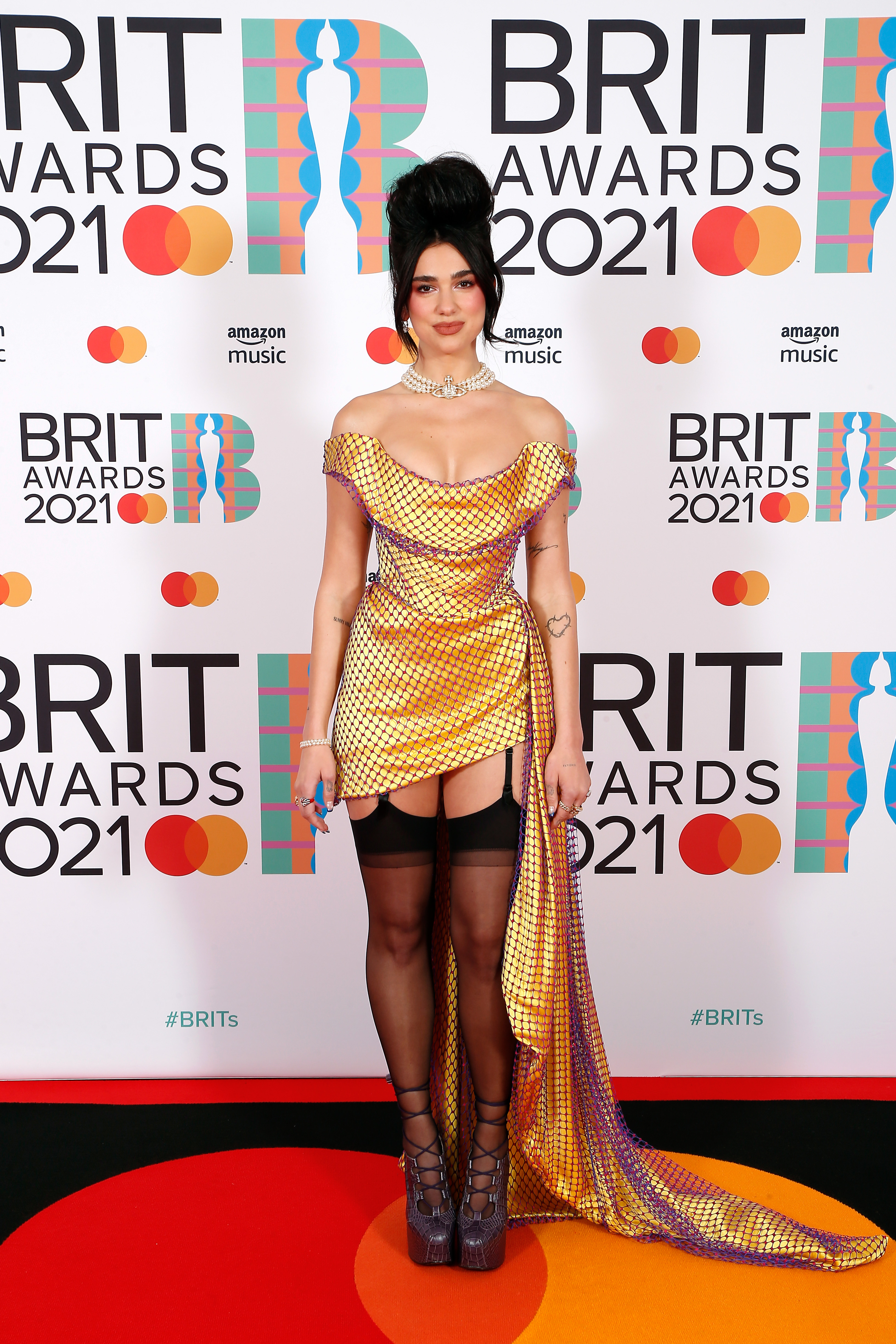 Dua Lipa no tapete vermelho do BRIT Awards 2021. Ela está com um leve sorriso sem mostrar os dentes, usando um vestido dourado com uma rede de furinhos roxos por cima, uma meia-calça preta e um sapato de salto. Também está com colar de pérolas e cabelo preso em um coque alto bem volumoso.