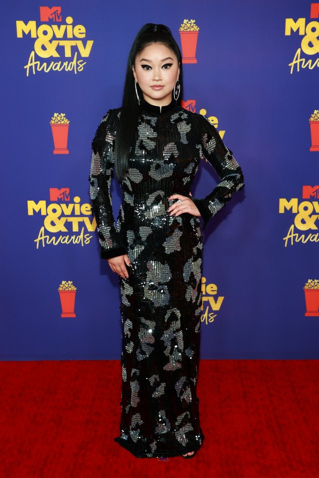 Atriz Lana Condor no MTV Movie & TV Awards, usando vestido longo e preto com detalhes em prata, posando com uma mão na cintura e expressão sorridente no red carpet da premiação