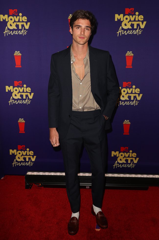 Jacob Elordi no MTv Movie & TV Awards, posando com expressão séria no red carpet da premiação usando terno preto e camisa levemente marrom.