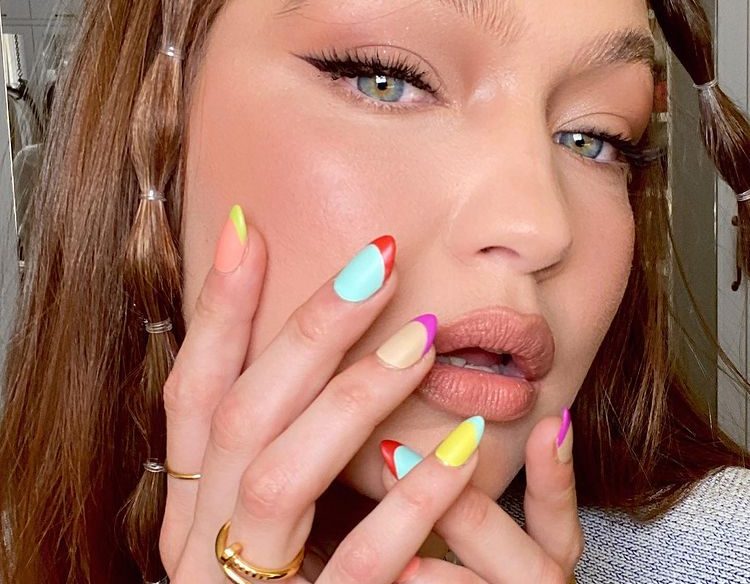 Gigi Hadid com nail art colorida Gigi Hadid com nail art de francesinha colorida, posando com as mãos no rosto para evidenciar as unhas, suas unhas estão com esmalte azul, amarelo, laranja, roxo e rosa. Sua expressão é seria. Nos cabelos ela tem duas mechas com bubble hair.