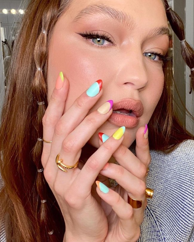 Gigi Hadid com nail art de francesinha colorida, posando com as mãos no rosto para evidenciar as unhas, suas unhas estão com esmalte azul, amarelo, laranja, roxo e rosa. Sua expressão é seria. Nos cabelos ela tem duas mechas com bubble hair.