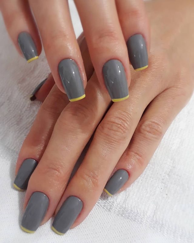 Foto mostrando uma mão com os dedos dobrados para evidenciar as unhas pintadas em francesinha colorida amarela com base cinza