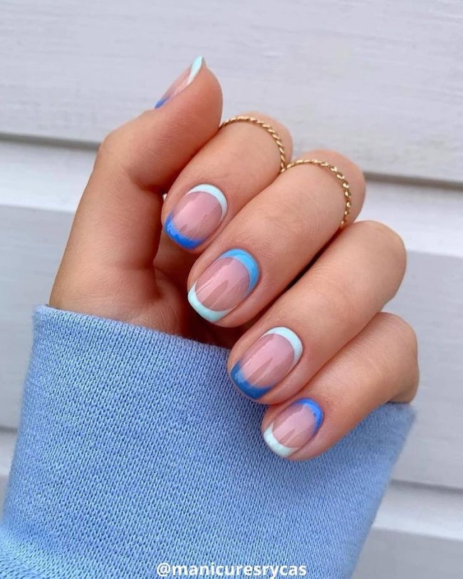 Foto mostrando uma mão com os dedos dobrados para evidenciar as unhas pintadas em francesinha colorida com a ponta e o começo da unha pintados com detalhes azul.