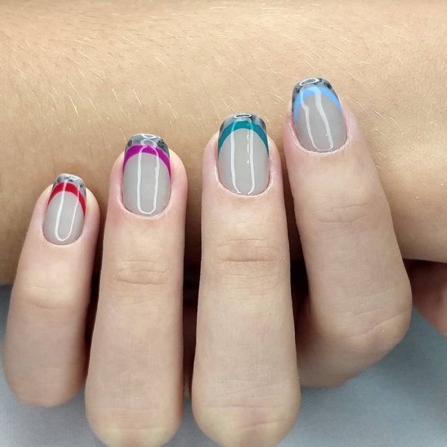Foto mostrando uma mão com os dedos dobrados para evidenciar as unhas pintadas em francesinha colorida; com base cinza e linhas coloridas, em roxo, vermelho, cinza e verde.