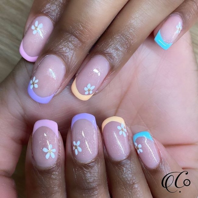 Duas mãos exibindo unhas com francesinha colorida nas cores lilás, azul e laranja. E florzinha.