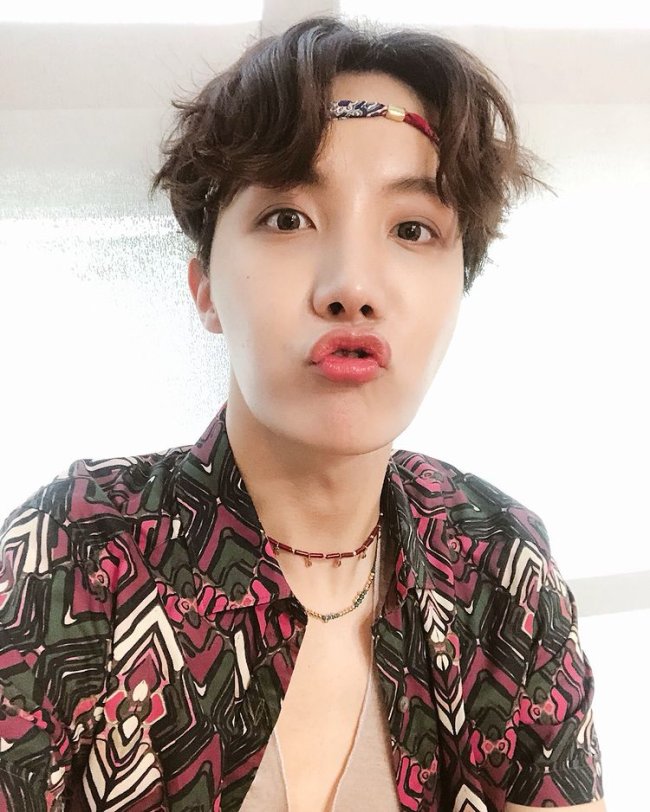 J-Hope, do grupo de k-pop BTS fazendo biquinho para foto, usando elástico na testa, colares e camisa estampada nos tons vermelho, verde, branco e preto.