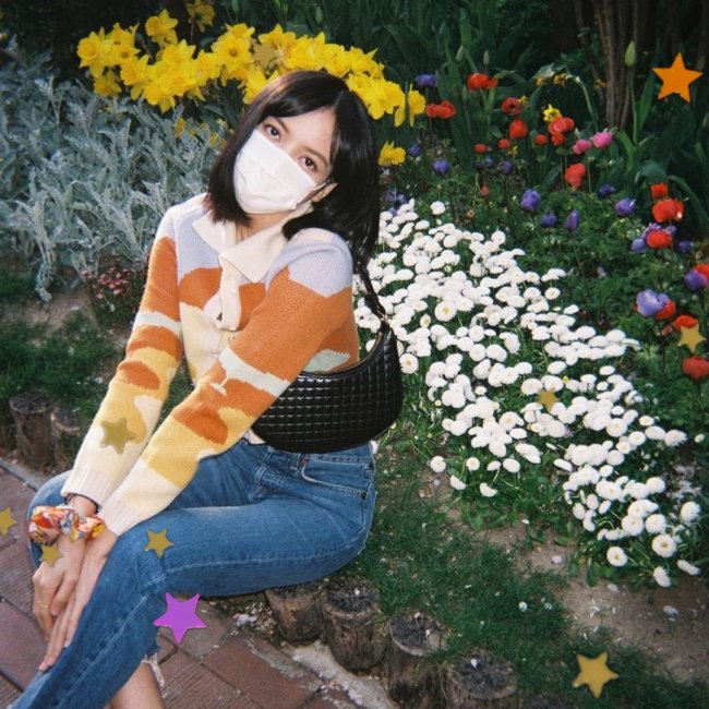 LISA BLACKPINK KPOP Lisa, integrante do grupo Blackpink, sentada perto de flores brancas, vermelhas e amarelas. Usando camisa listrada azul clara, laranja e amarela. Calça jeans, e bolsa baguete preta. Não conseguimos ver sua expressão pois Lisa está usando uma máscara de proteção branca.
