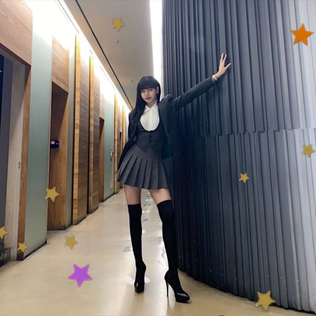 LISA BLACKPINK Lisa, integrante do grupo Blackpink, posando para foto com uma das mãos apoiada na parede, usando bota over the knee, saia colegial cinza e com uma expressão séria. Ela esta posando em um corredor.